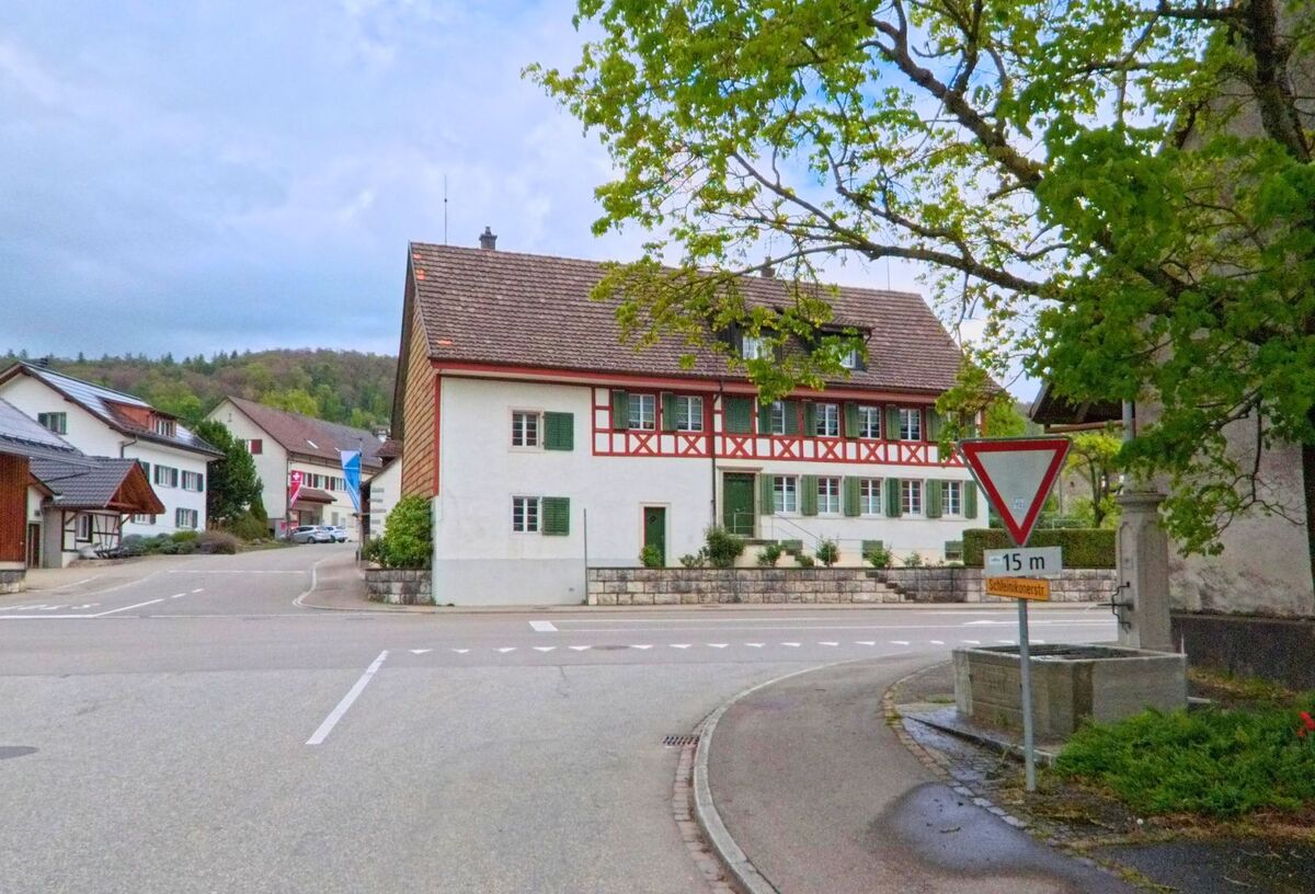 Im Zentrum von Oberweningen, PLZ 8165 im Kanton Zürich, steht ein historisches Fachwerkhaus an einer verkehrsberuhigten Kreuzung mit Brunnen.