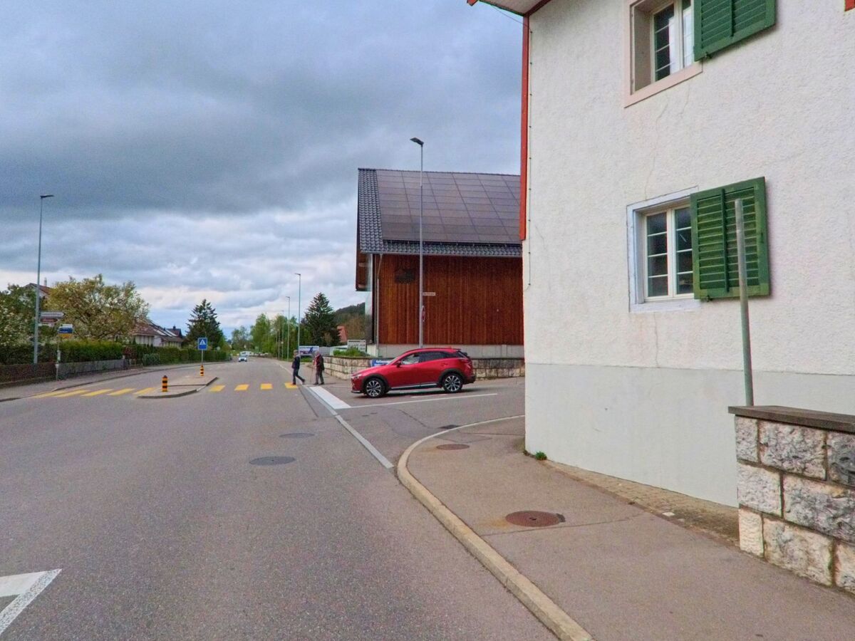 In Oberweningen, PLZ 8165 im Kanton Zürich, überquert eine Person einen gelb markierten Fussgängerstreifen bei einem Holzgebäude mit Solardach.