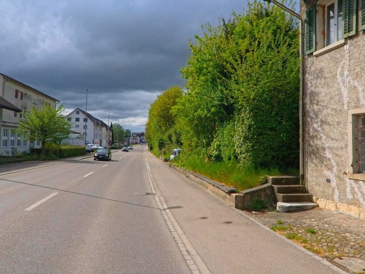 Bei PLZ 8165 in Oberweningen im Kanton Zürich verläuft eine stark befahrene Strasse zwischen Wohnhäusern und dichter Begrünung unter dunklen Regenwolken.