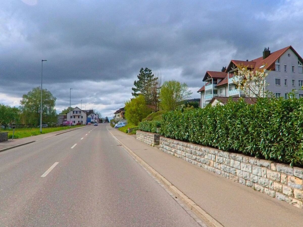  In Oberweningen, PLZ 8165 im Kanton Zürich, verläuft eine Strasse mit breitem Gehweg entlang von Mehrfamilienhäusern und gepflegten Hecken unter bewölktem Himmel.