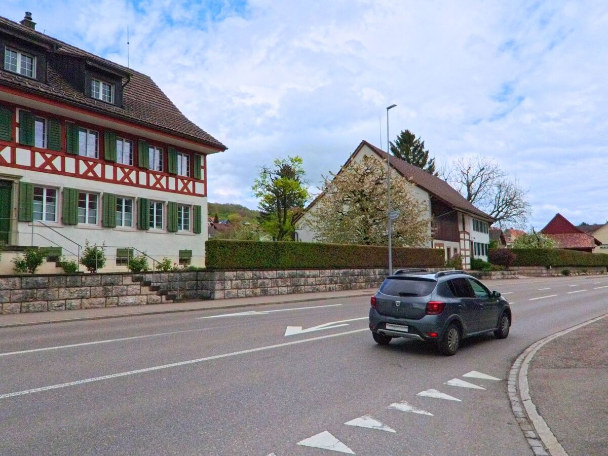 In Oberweningen, PLZ 8165 im Kanton Zürich, passiert ein Fahrzeug ein traditionelles Fachwerkhaus mit grün gestrichenen Fensterläden an einer breiten Dorfstrasse. Bildtitel: oberweningen8165verkehr_fachwerkhaus