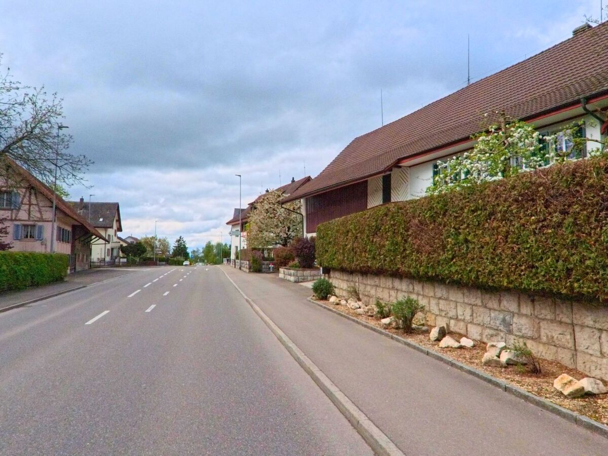 In Oberweningen, PLZ 8165 im Kanton Zürich, verläuft eine ruhige Strasse zwischen gepflegten Wohnhäusern mit Vorgärten und Hecken.