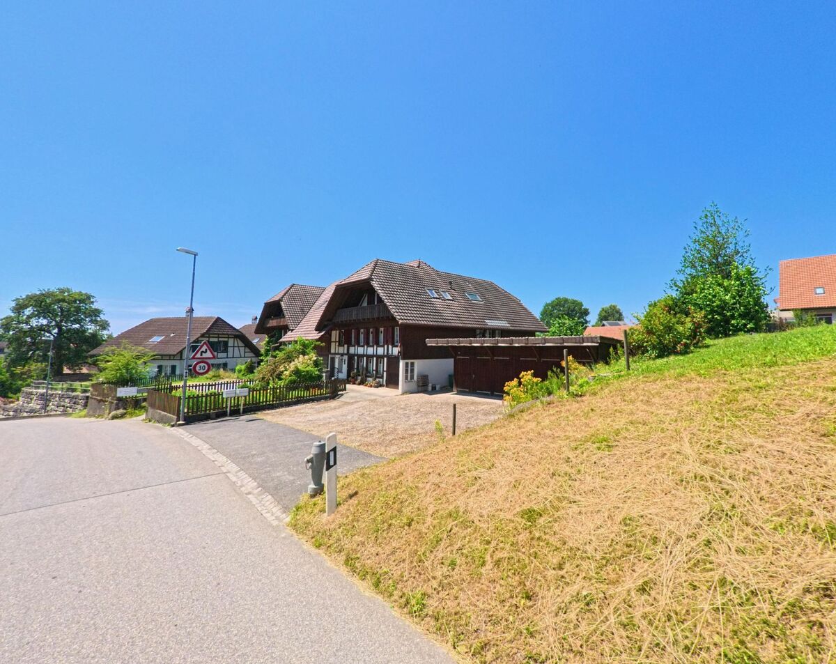 In Oberwil bei Büren (3298, Kanton Bern) führt eine Straße an traditionellen Fachwerkhäusern und grünen Flächen vorbei, während im Hintergrund Bäume und gepflegte Gärten sichtbar sind.