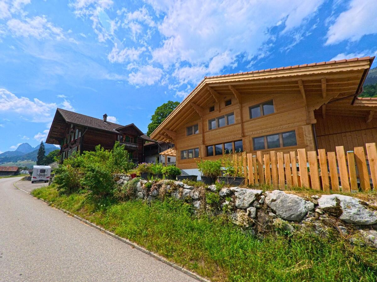 In Oberwil im Simmental (3765, Kanton Bern) stehen ein traditionelles Bauernhaus und ein modernes Holzhaus nebeneinander an einer geschwungenen Strasse mit Blick auf die Berge.