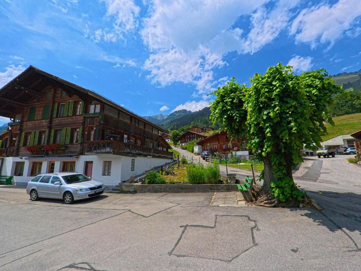 In Oberwil im Simmental (3765, Kanton Bern) steht ein mächtiges Holzhaus mit grünen Fensterläden neben einer Baumgruppe mit Sitzbank an einem kleinen Platz.