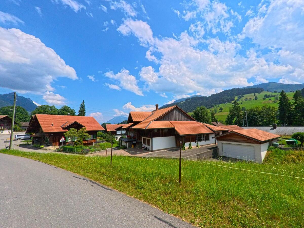 In Oberwil im Simmental (3765, Kanton Bern) reihen sich Einfamilienhäuser mit roten Ziegeldächern entlang einer leicht ansteigenden Strasse mit Blick auf bewaldete Hügel.