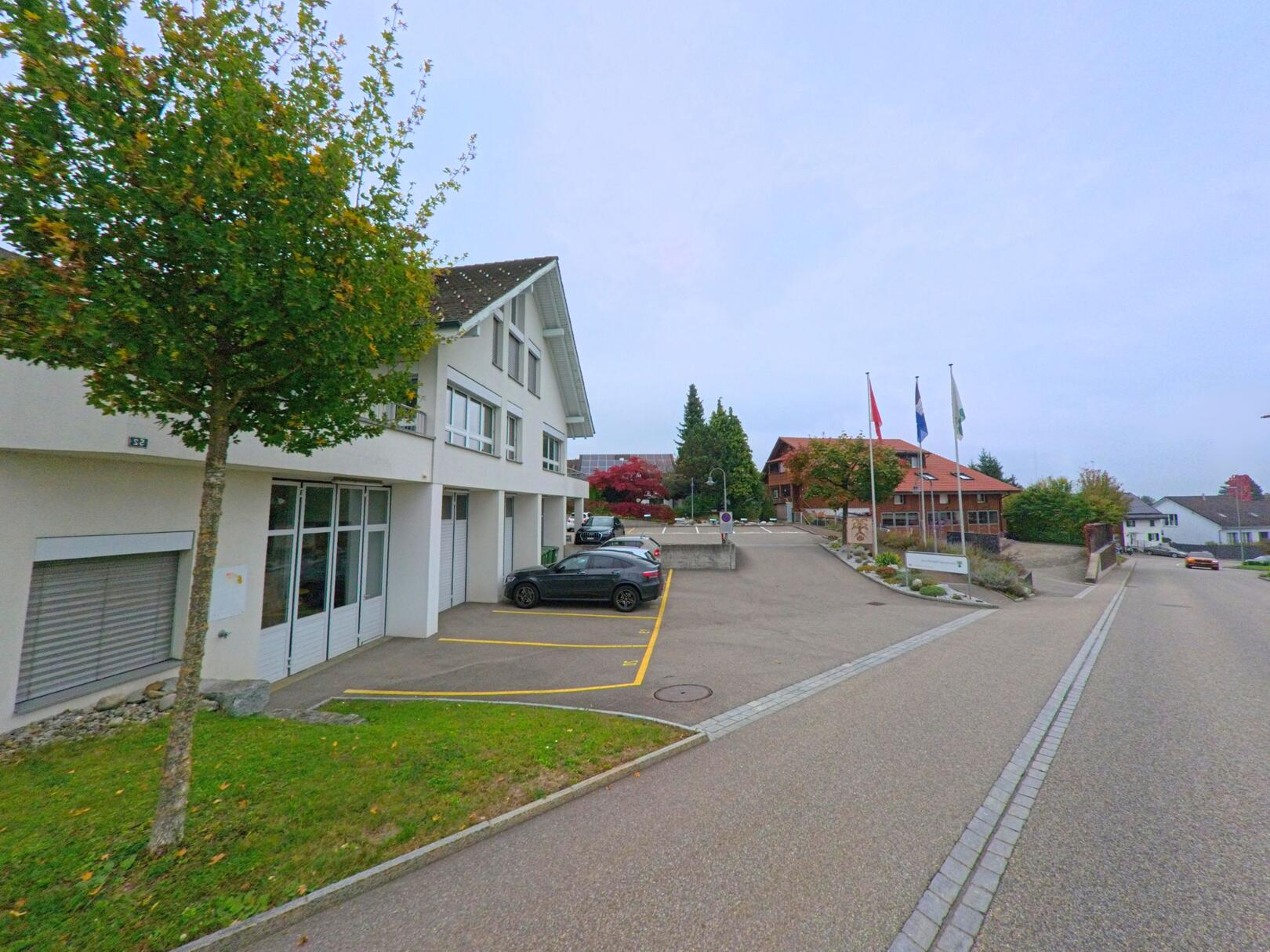 Vor dem Gemeindehaus in Oberwil-Lieli (8966, Kanton Aargau) liegt eine ruhige Strasse mit modernen und traditionellen Gebäuden sowie wehenden Fahnen.