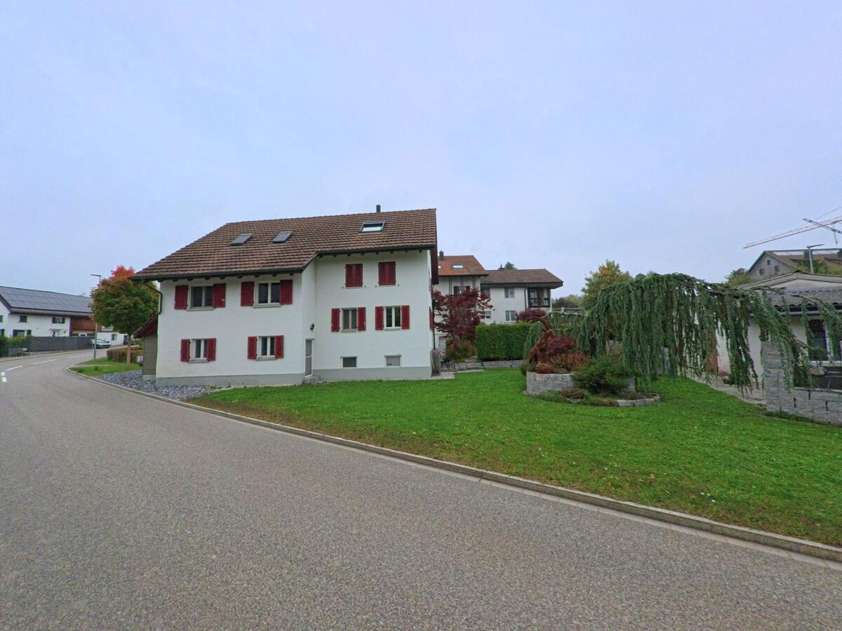 Ein weiss gestrichenes Wohnhaus mit roten Fensterläden und gepflegtem Garten steht an einer leicht gebogenen Strasse in Oberwil-Lieli (8966, Kanton Aargau).