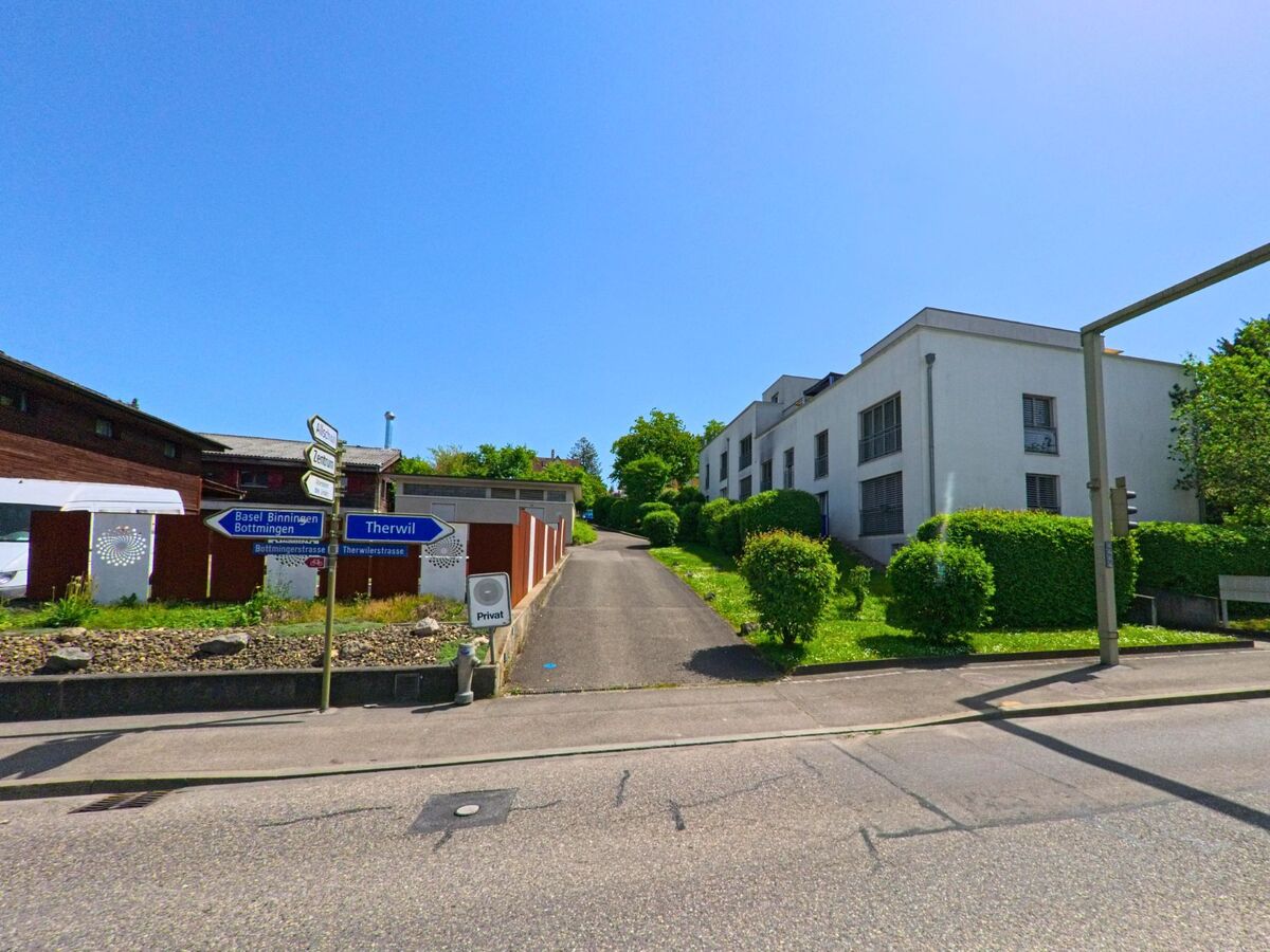 In Oberwil (PLZ 4104, Kanton Basel-Landschaft) zweigt eine private Zufahrtsstrasse mit modernen Wohnbauten von der Therwilerstrasse ab, flankiert von Wegweisern in Richtung Therwil und Basel.