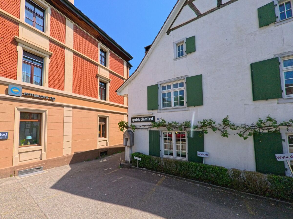 In Oberwil (PLZ 4104, Kanton Basel-Landschaft) liegen ein historisches Fachwerkhaus mit gruenen Fensterlaeden und ein Backsteingebaeude direkt nebeneinander im Zentrum.