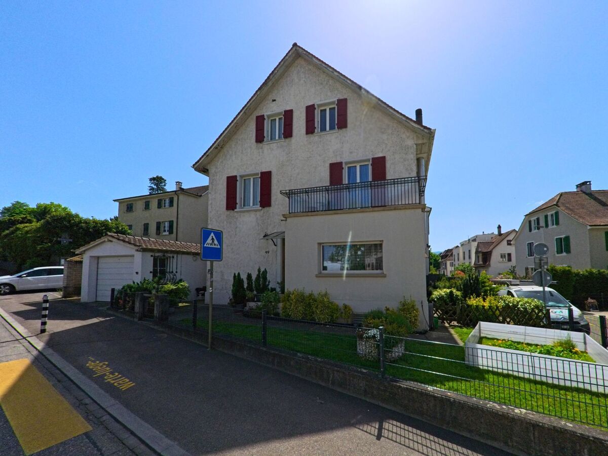 In Oberwil (4104, Kanton Basel-Landschaft) steht ein gepflegtes Einfamilienhaus mit roten Fensterläden und Vorgarten direkt an einem Fussgängerstreifen.