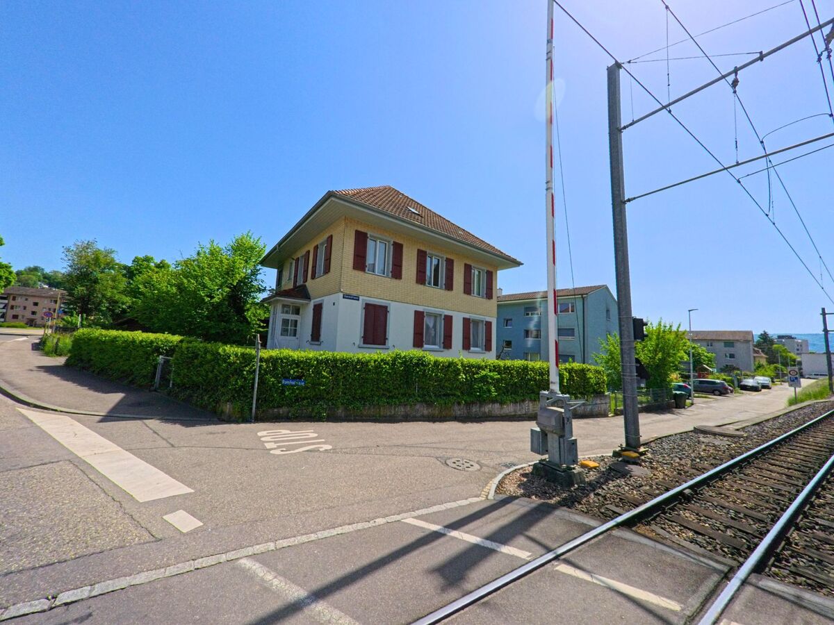 In Oberwil (4104, Kanton Basel-Landschaft) steht ein Wohnhaus mit roten Fensterlaeden direkt an einem beschrankten Bahnuebergang mit Gleisen und Oberleitungen.