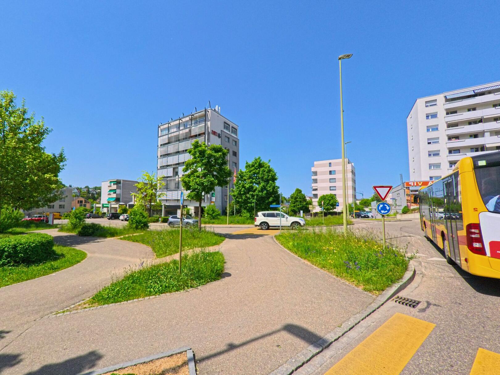 In Oberwil (4104 im Kanton Basel-Landschaft) zeigt sich ein moderner Kreisverkehr mit Wohnbauten und Bushaltestelle an einem sonnigen Tag.
