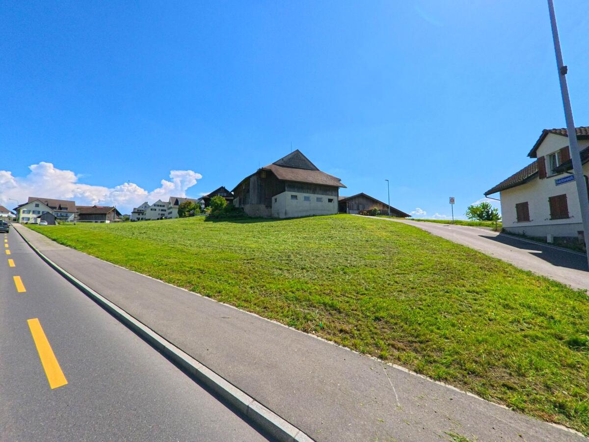  In Obfelden, PLZ 8912 im Kanton Zürich, steigt eine Nebenstrasse von einer Hauptstrasse zu einem Hügel mit landwirtschaftlichen Gebäuden und Wohnhäusern an.