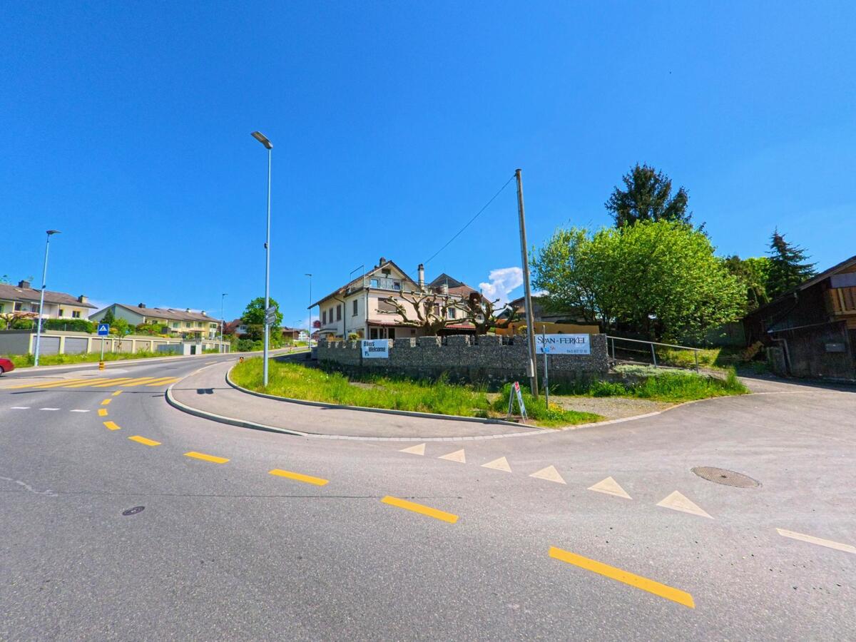 In Obfelden, PLZ 8912 im Kanton Zürich, verzweigen sich mehrere Strassen an einem kleinen Platz mit Einfamilienhäusern, Werbeschildern und Baumgruppen.