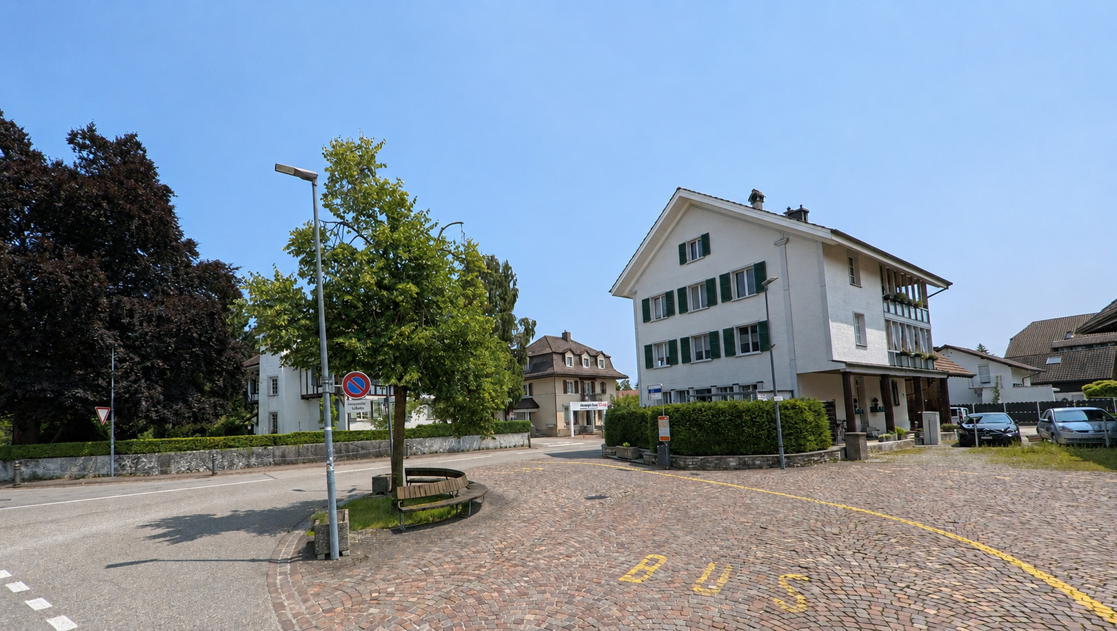 An einer Bushaltestelle in Oekingen (4566, Kanton Solothurn) stehen mehrere Wohnhäuser mit grünen Fensterläden rund um einen gepflasterten Vorplatz. Bildtitel: oekingen_4566_bushaltestelle_mit_gepflastertem_vorplatz.