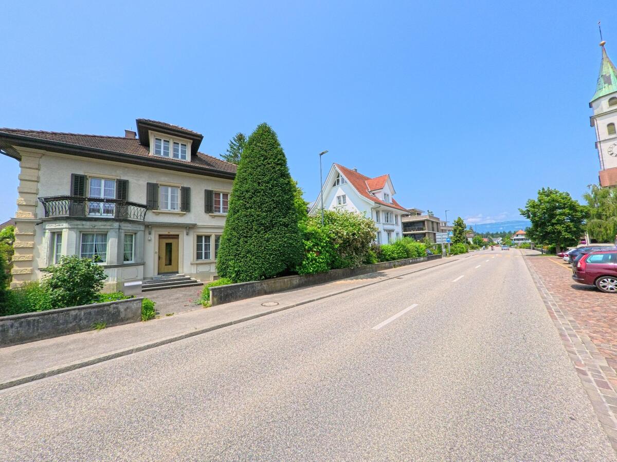 Entlang einer breiten Dorfstrasse in Oekingen (4566, Kanton Solothurn) stehen gepflegte Wohnhäuser mit Gärten, während am rechten Bildrand ein Kirchturm sichtbar ist..jpg