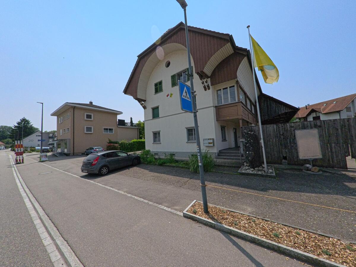 An einer Dorfstrasse in Oekingen (4566, Kanton Solothurn) steht ein markantes Gebäude mit geschwungenem Giebel, grünen Fensterläden und einer Fahne am Vorplatz.