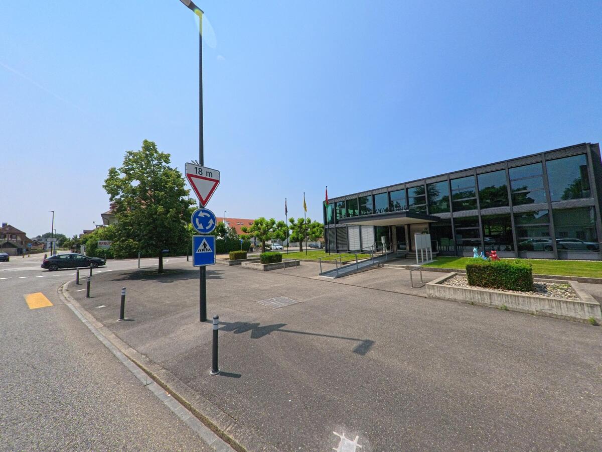 Neben einem kleinen Kreisverkehr steht in Oekingen (4566, Kanton Solothurn) ein modernes öffentliches Gebäude mit grosser Glasfassade, Fahnen und gepflastertem Vorplatz..jpg