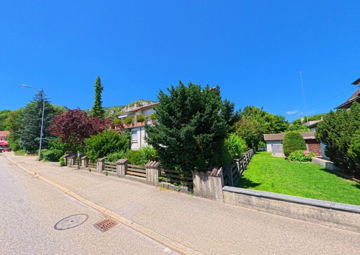 In Oensingen (4702, Kanton Solothurn) säumen Einfamilienhäuser mit dichtem Gartenbewuchs eine ruhige Strasse am Fuss eines bewaldeten Hangs.