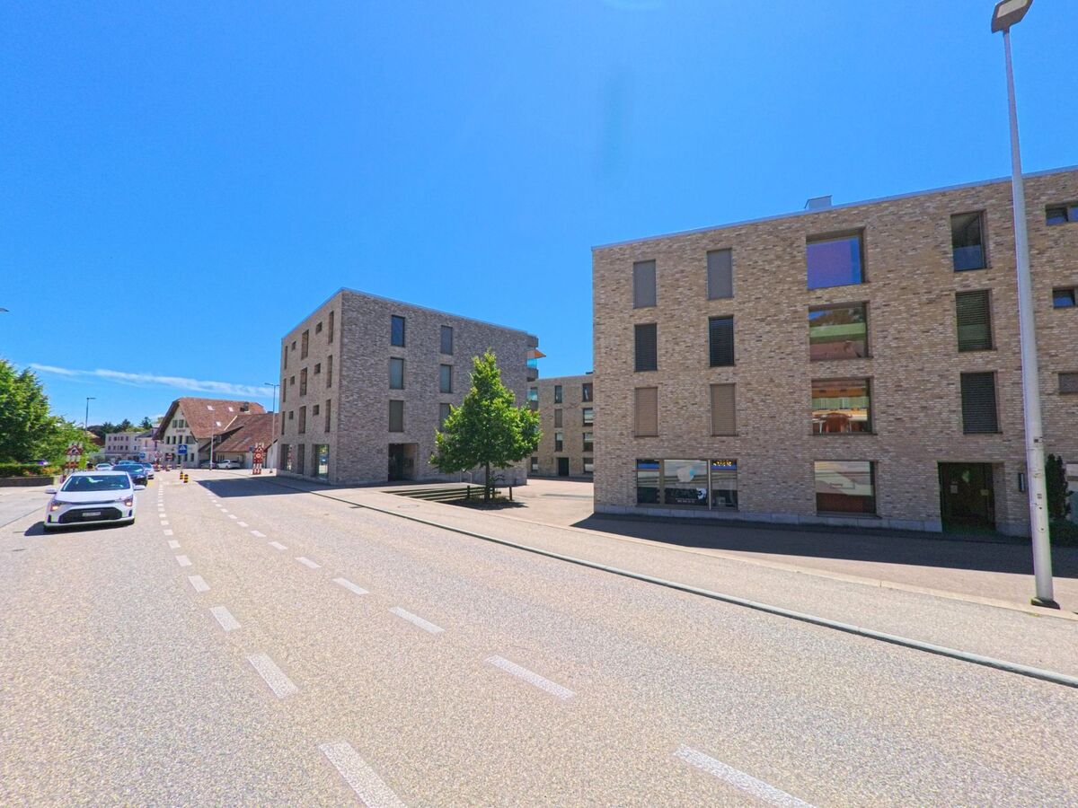 In Oensingen (4702, Kanton Solothurn) säumen moderne Wohn- und Geschäftsbauten mit heller Klinkerfassade eine mehrspurige Strasse mit Markierungen für den Verkehr.