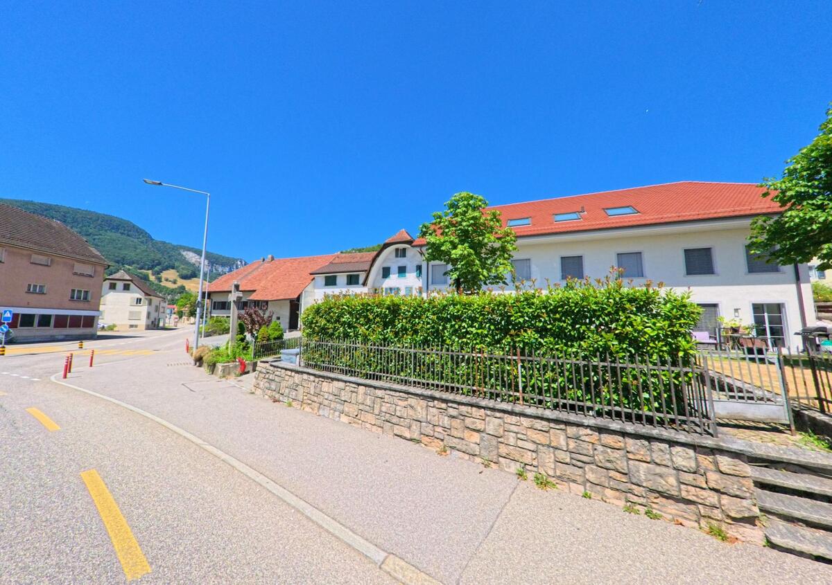 In Oensingen (4702, Kanton Solothurn) verläuft eine Strasse mit modernen und traditionellen Wohnhäusern entlang einer bepflanzten Mauer, im Hintergrund erheben sich bewaldete Hügel.