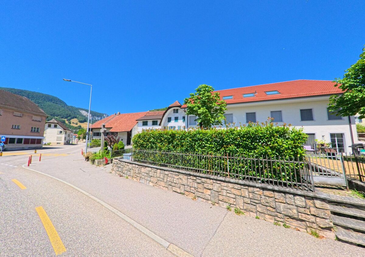 In Oensingen (4702, Kanton Solothurn) verläuft eine Strasse mit modernen und traditionellen Wohnhäusern entlang einer bepflanzten Mauer, im Hintergrund erheben sich bewaldete Hügel.