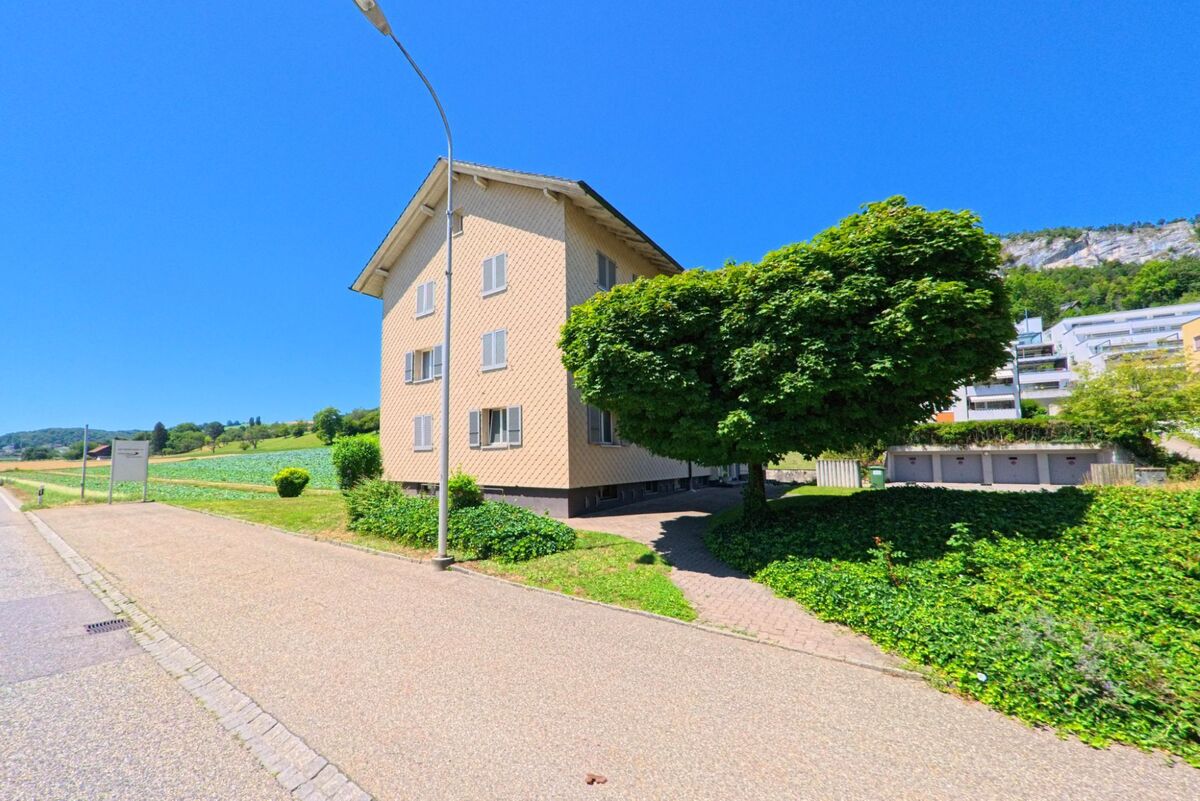 In Oensingen (4702, Kanton Solothurn) steht ein Wohnhaus mit Fensterläden am Rand eines Feldes, im Hintergrund sind moderne Wohnanlagen und ein Felsmassiv sichtbar.