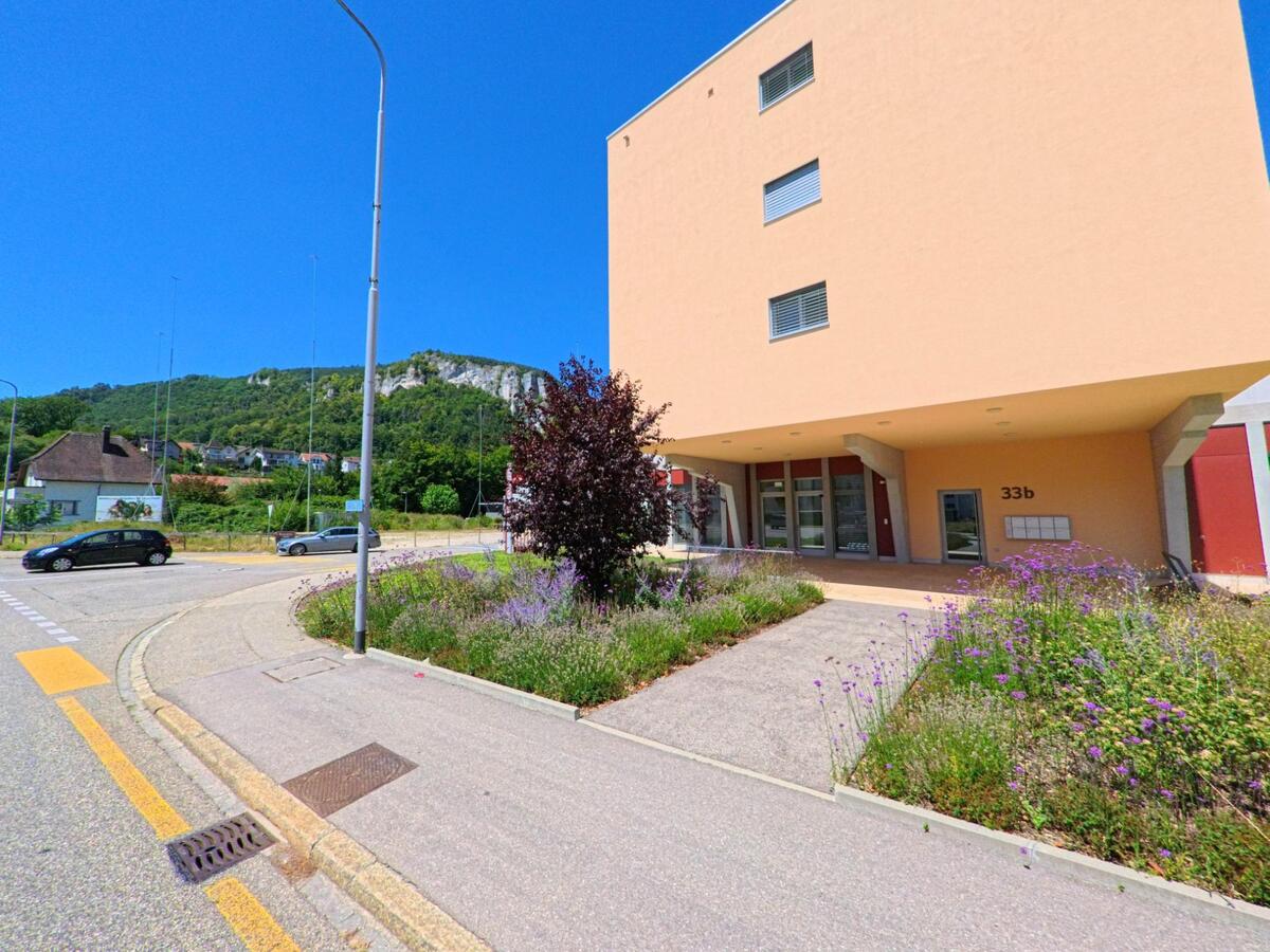 In Oensingen (4702, Kanton Solothurn) liegt ein modernes Wohnhaus mit bepflanztem Vorbereich an einem Verkehrskreisel, im Hintergrund erhebt sich ein bewaldeter Felsrücken.