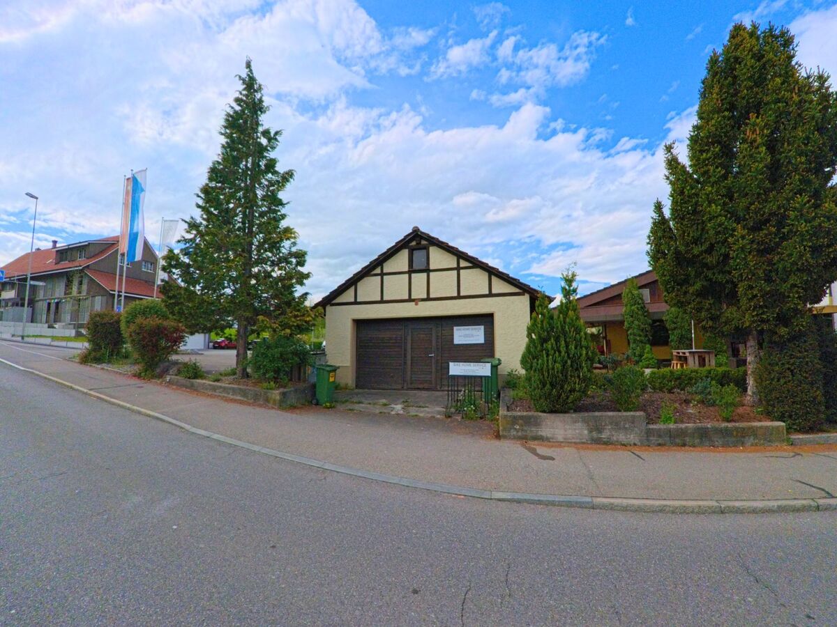 In Oetwil am See im Kanton Zürich (PLZ 8618) steht ein kleines Gebäude mit Fachwerkelementen und dunkelbraunen Toren an einer ruhig verlaufenden Strasse.