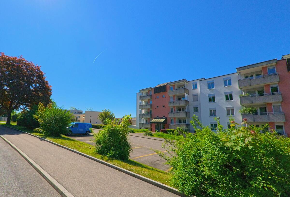 In Oetwil an der Limmat im PLZ-Gebiet 8955 des Kantons Zürich steht ein farblich akzentuiertes Mehrfamilienhaus mit Balkonen neben einem Parkplatz und gepflegten Grünflächen.