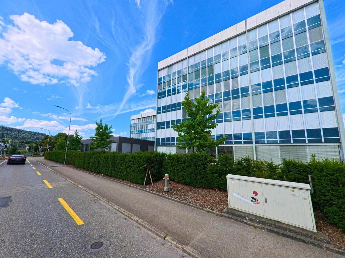 Entlang einer Strasse in Oftringen (4665, Kanton Aargau) erstreckt sich ein modernes Buero- und Gewerbegebaeude mit grosszuegiger Glasfassade hinter einer gepflegten Hecke.