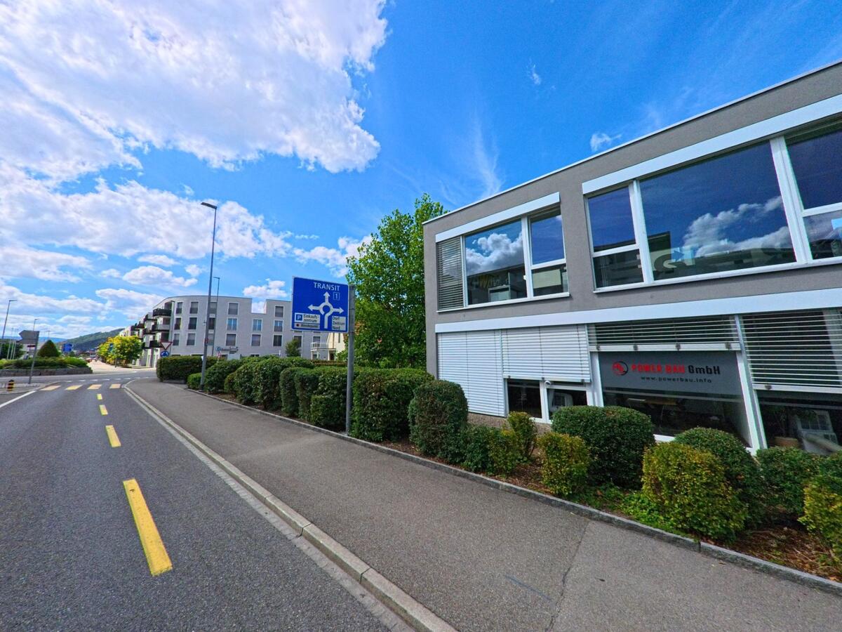 An einer breiten Strasse in Oftringen (4665, Kanton Aargau) liegt das Gewerbegebaeude der Power Bau GmbH, waehrend ein Verkehrszeichen auf eine bevorstehende Kreisverkehrsregelung hinweist.