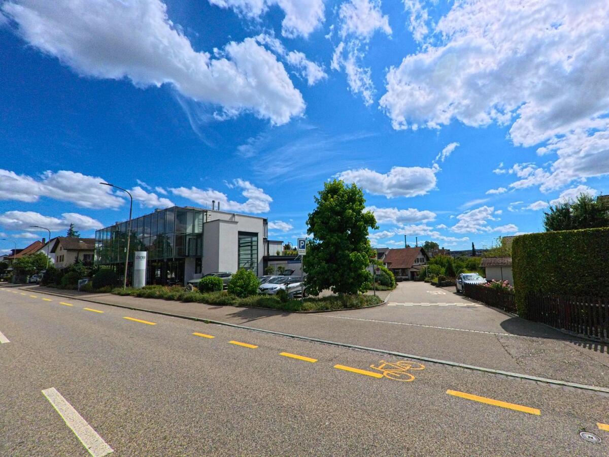 Ein modernes Glasgebaeude mit angeschlossenem Parkplatz steht in Oftringen (4665, Kanton Aargau) an einer verkehrsberuhigten Strassenecke mit angrenzendem Wohnquartier.