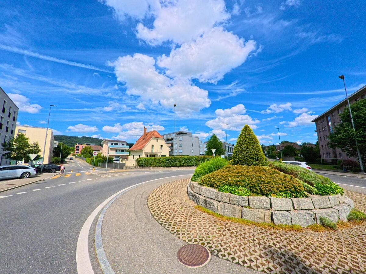 Ein begruenter Kreisverkehr mit gepflegter Bepflanzung und Steinmauer praegt das Ortsbild von Oftringen (4665, Kanton Aargau), umgeben von Wohnhaeusern und einer verkehrsberuhigten Strasse.