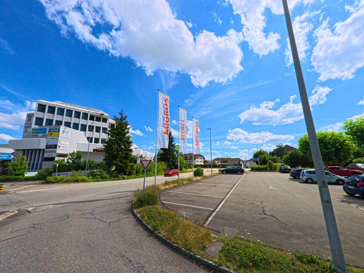 Neben einem weitlaeufigen Parkplatz in Oftringen (4665, Kanton Aargau) ragen mehrere Migros-Fahnen in die Hoehe, waehrend im Hintergrund ein markantes Geschaeftsgebaeude mit zahlreichen Firmenschildern steht.
