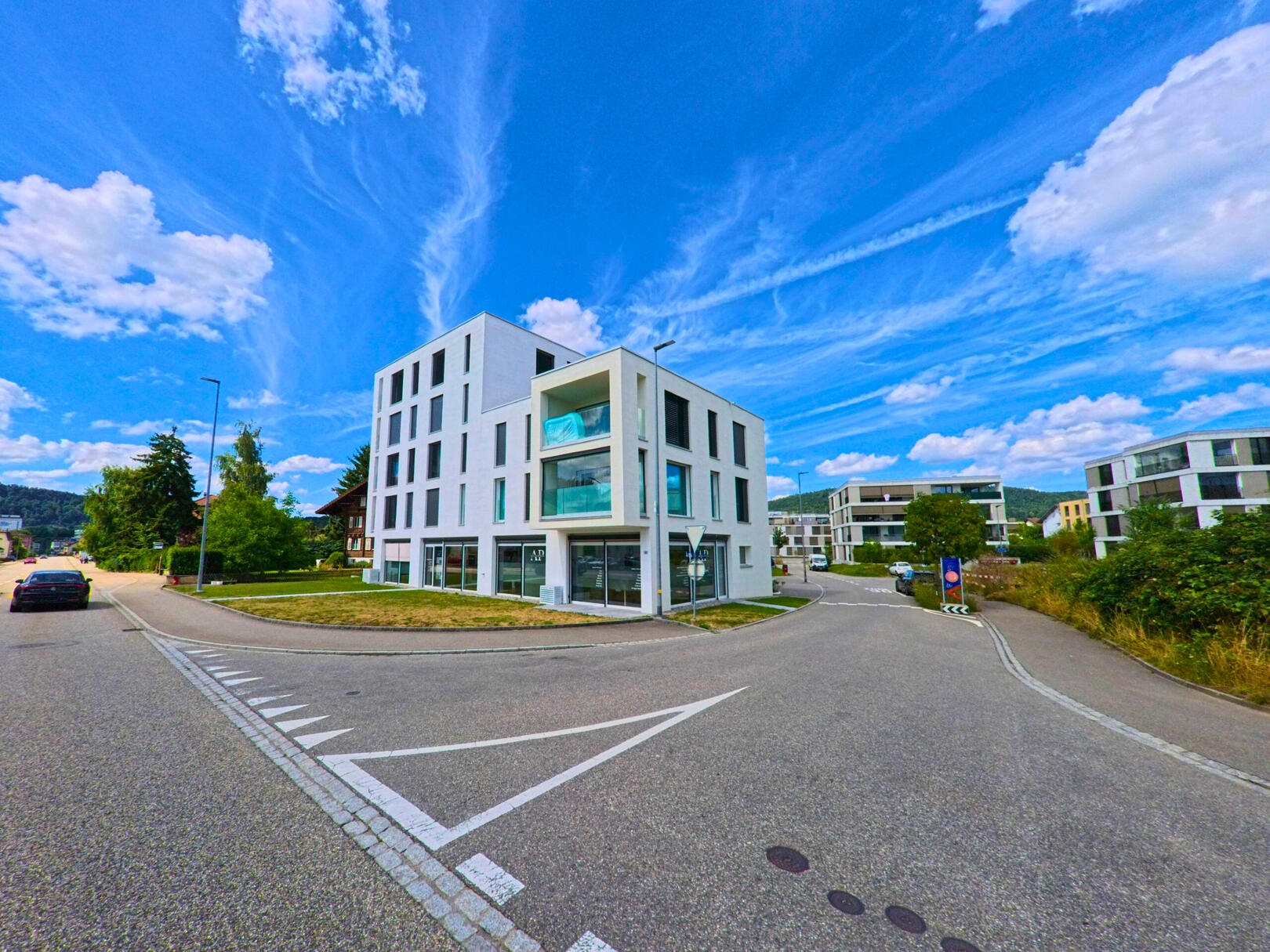 In Oftringen (4665, Kanton Aargau) steht ein modernes, mehrstöckiges Wohngebäude an einer ruhig gelegenen Strassenkreuzung mit weiteren Neubauten im Hintergrund.