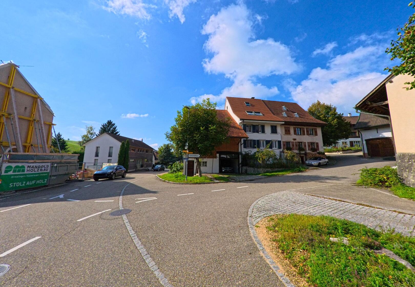 In Olsberg (4305, Kanton Aargau) zweigt eine Strasse an einer kleinen Kreuzung mit Wohnhäusern und Baustelle bei sonnigem Wetter ab.