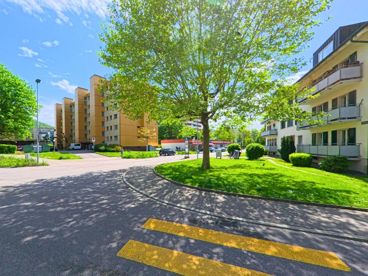 In Olten (4600, Kanton Solothurn) fuehrt ein Fussgaengerstreifen durch ein Wohnquartier mit mehrstöckigen Wohnhaeusern und gepflegter Gruenflaeche unter schattenspendenden Baeumen.