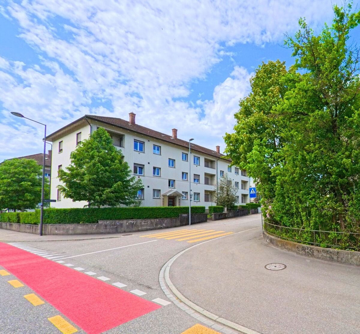 In Olten (4600, Kanton Solothurn) windet sich eine Wohnstrasse mit Fussgaengerstreifen und Radweg entlang eines Baumgürtels und mehrgeschossiger Wohnbauten.