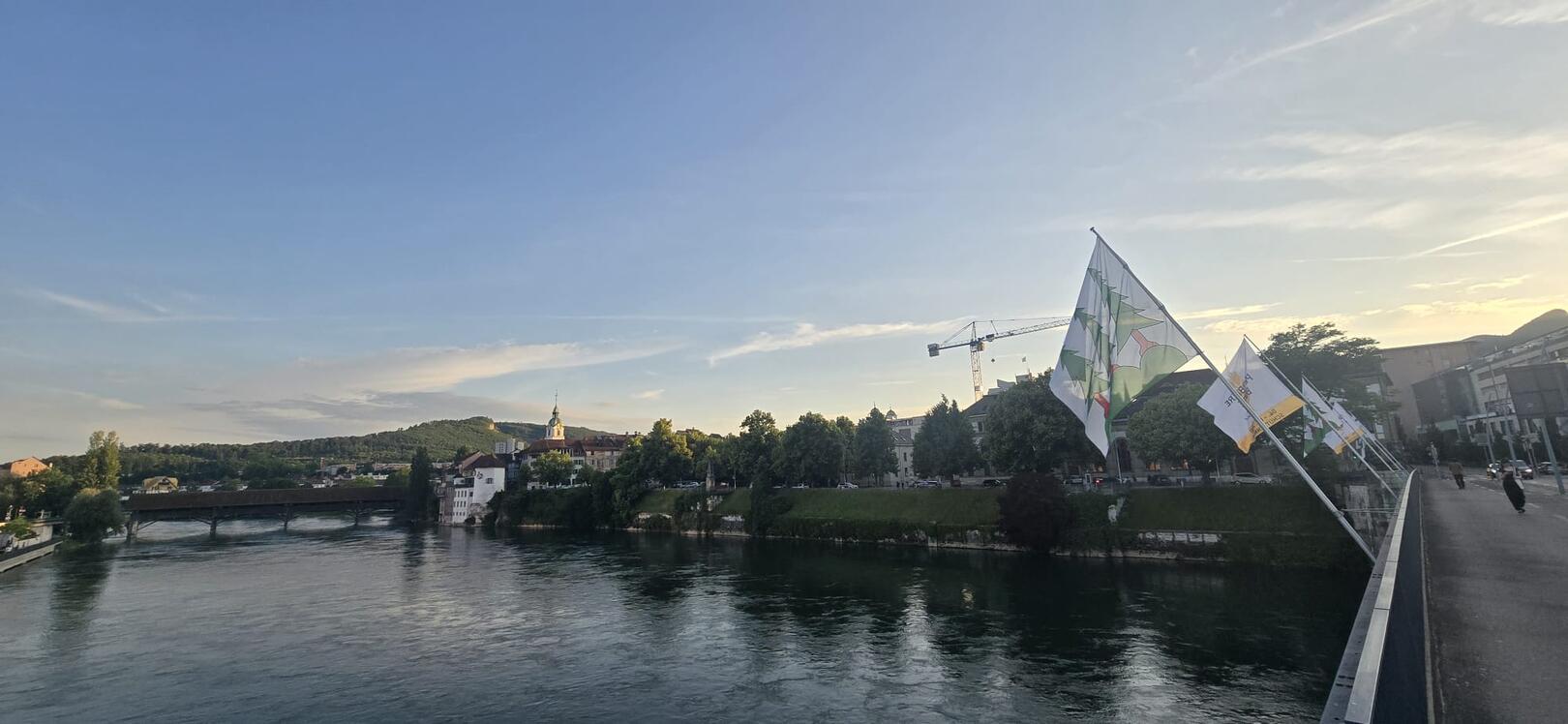 Blick über die Aare auf die Altstadt von Olten bei Abendstimmung, mit Brücke, Uferpromenade und wehenden Fahnen unter klarem Himmel im Kanton Solothurn.