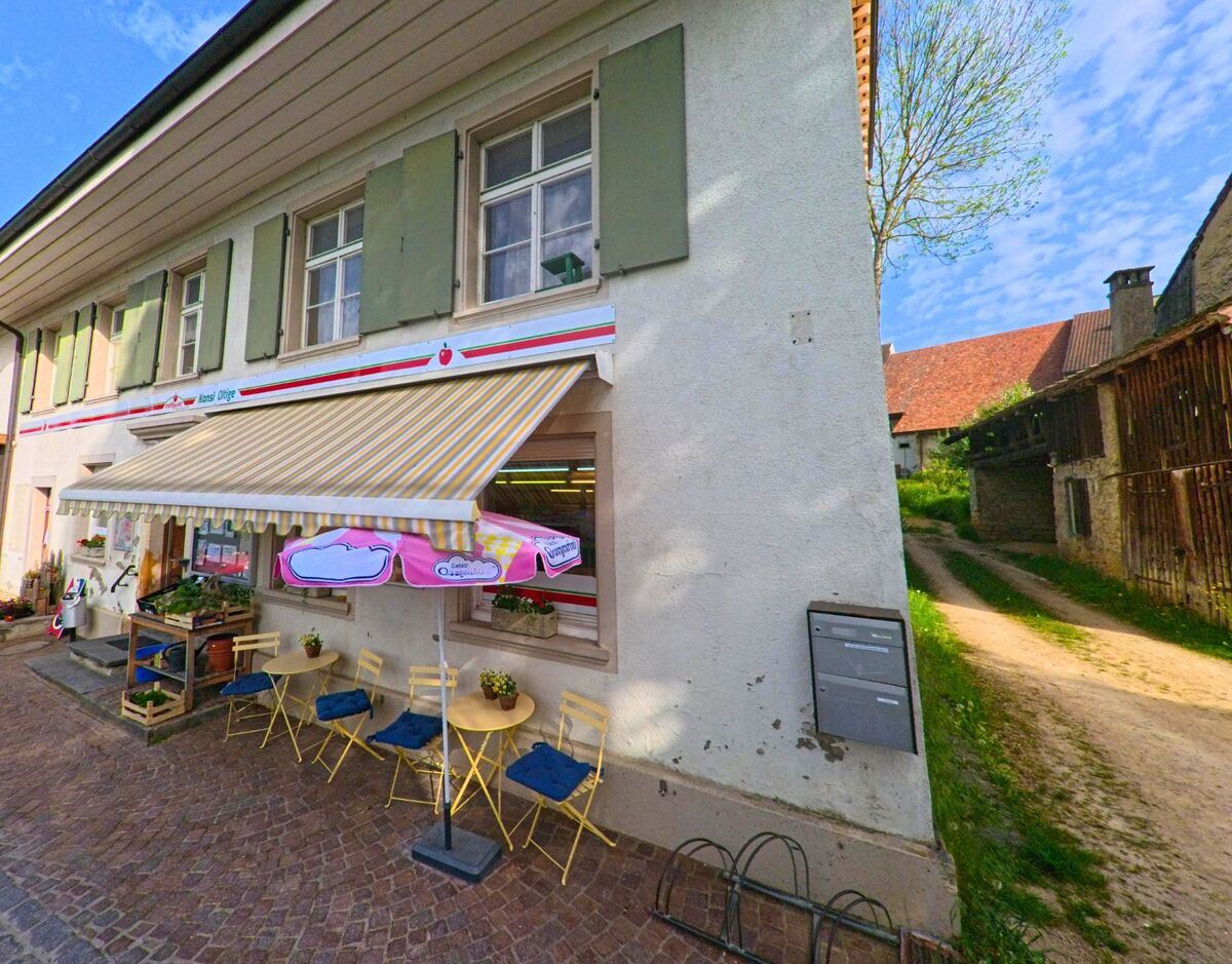In Oltingen (4494, Kanton Baselland) lädt ein kleiner Dorfladen mit Aussenbestuhlung, Markise und bunten Sonnenschirmen zum Verweilen ein.