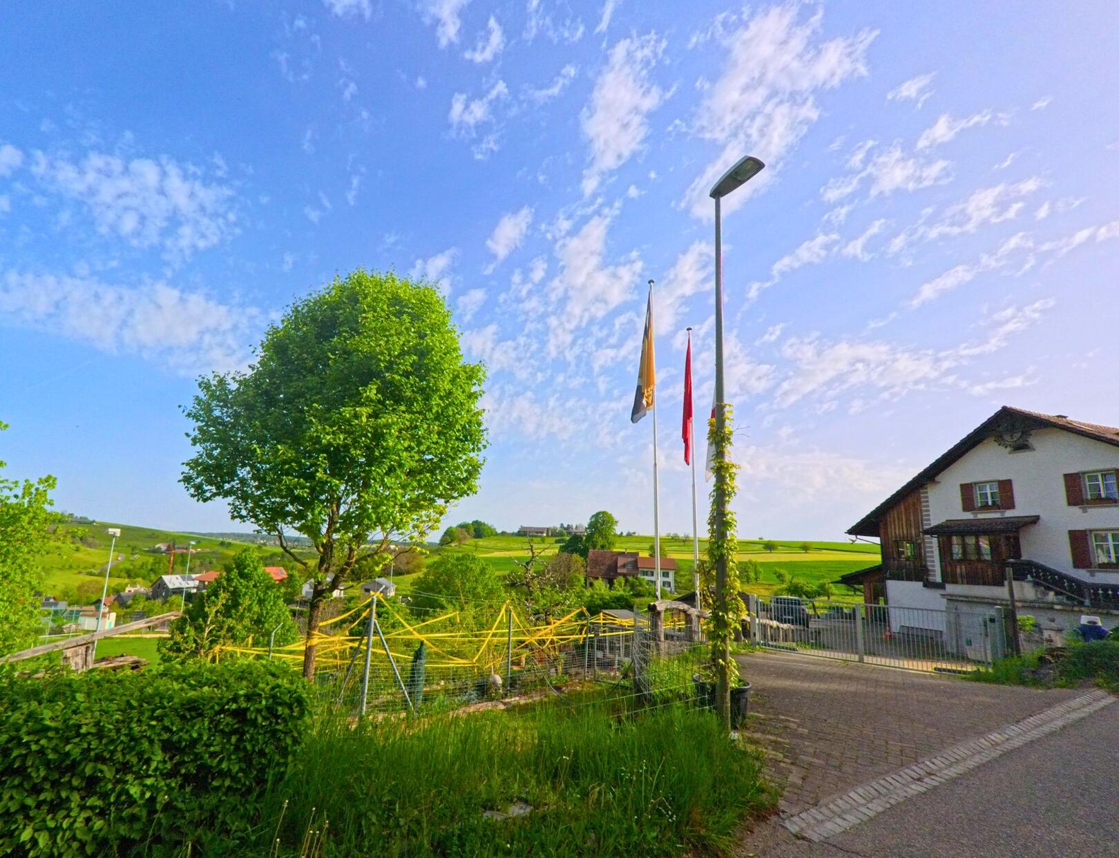 In Oltingen (4494, Kanton Baselland) wehen zwei Fahnen neben einem Wohnhaus, während im Hintergrund die grüne Hügellandschaft sichtbar ist.
