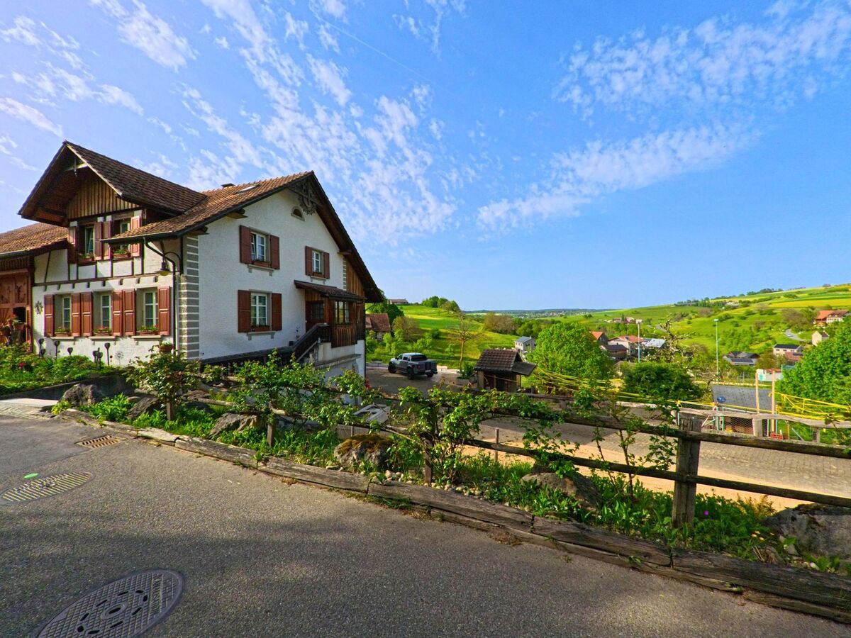 In Oltingen (4494, Kanton Baselland) steht ein traditionelles Wohnhaus mit roten Fensterläden oberhalb eines Tals mit weitem Blick über die hügelige Landschaft.