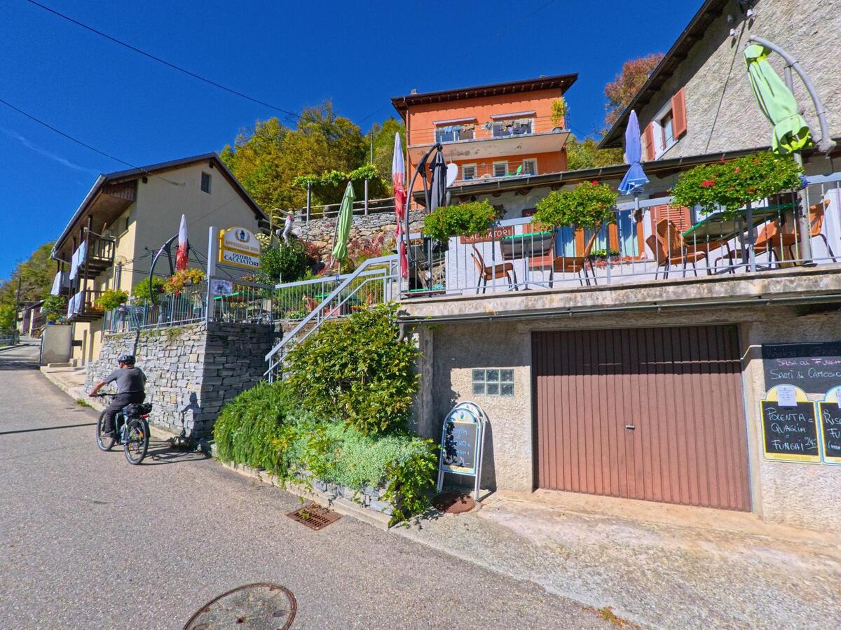 Ein Radfahrer fährt an einem Restaurant mit sonniger Terrasse in Onsernone (6661, Kanton Tessin) vorbei, das an einem Hang mit blumengeschmückten Balkonen liegt.