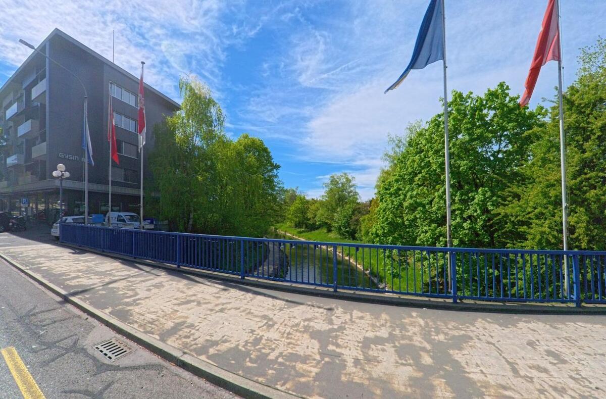 Eine Bruecke mit blauen Gelaendern ueberquert in Opfikon (8152, Kanton Zürich) einen von Baeumen gesaeumten Fluss, neben einem modernen Gebaeude mit Fahnen.