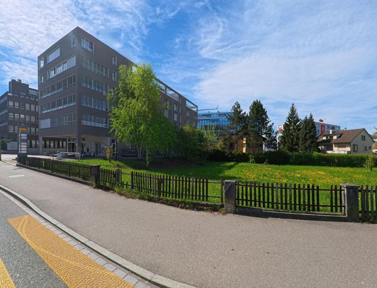 In Opfikon (8152, Kanton Zürich) steht ein modernes Buerohaus neben einer Gruenflaeche mit Holzzaun, hinter der sich Wohnhaeuser befinden.