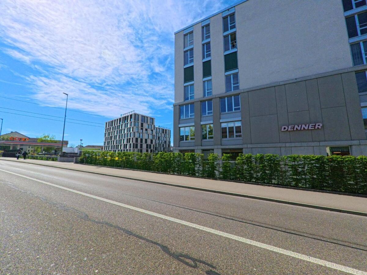 An einer Hauptstrasse in Opfikon (8152, Kanton Zürich) befinden sich ein Denner-Markt im Vordergrund und ein Coop-Gebaeude im Hintergrund neben modernen Buerohaeusern.