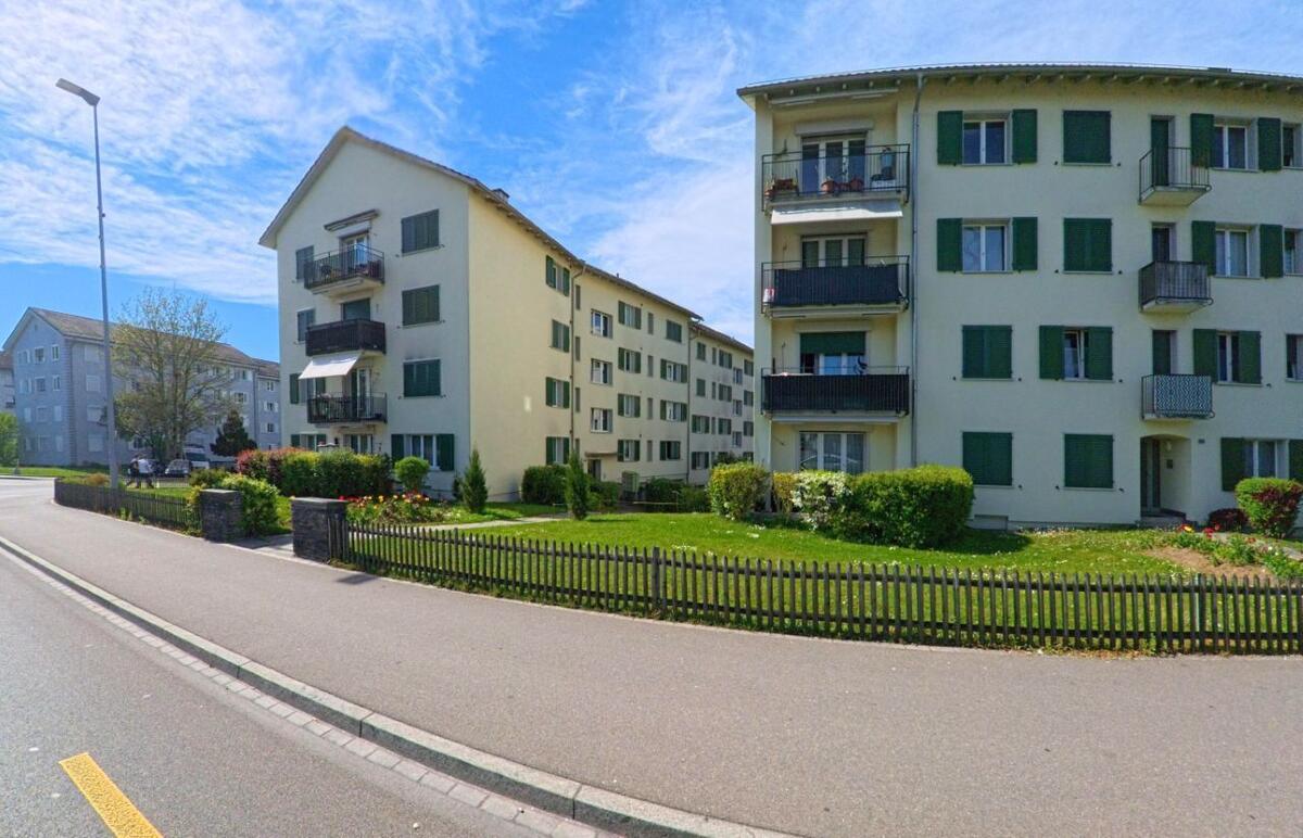 Mehrfamilienhaeuser mit gruener Fassade und Balkonen stehen in Opfikon (8152, Kanton Zürich) an einer Strasse, getrennt durch eine kleine Gartenflaeche mit Holzzaun.