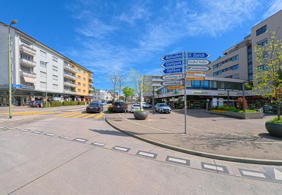 Im Zentrum von Opfikon (8152, Kanton Zürich) stehen Wegweiser vor Geschaefts- und Wohngebaeuden an einer Strasse mit Fussgaengerstreifen.